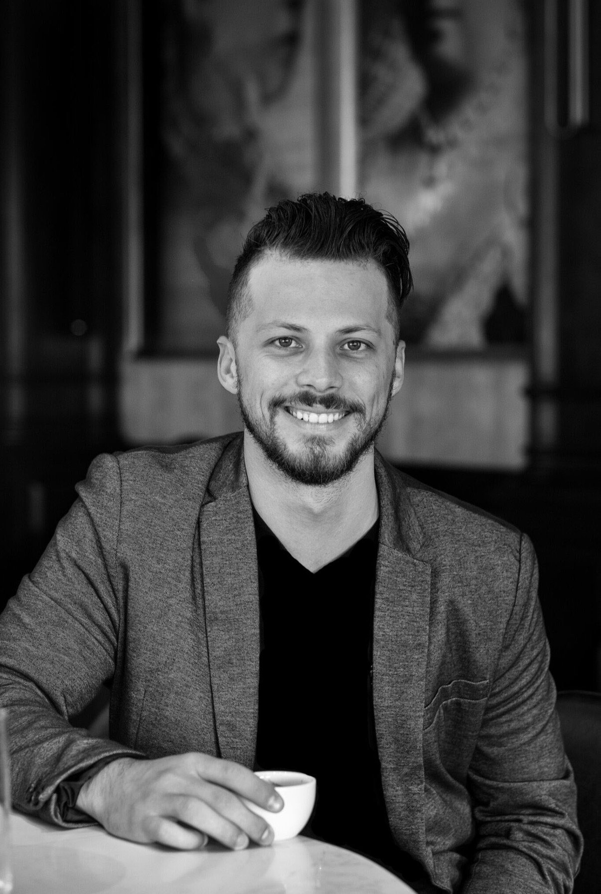 A black and white photo of a smiling man with a beard and styled hair, dressed in a blazer and black shirt, holding a small cup in his right hand, sitting at a table in a room with artwork in the background.
