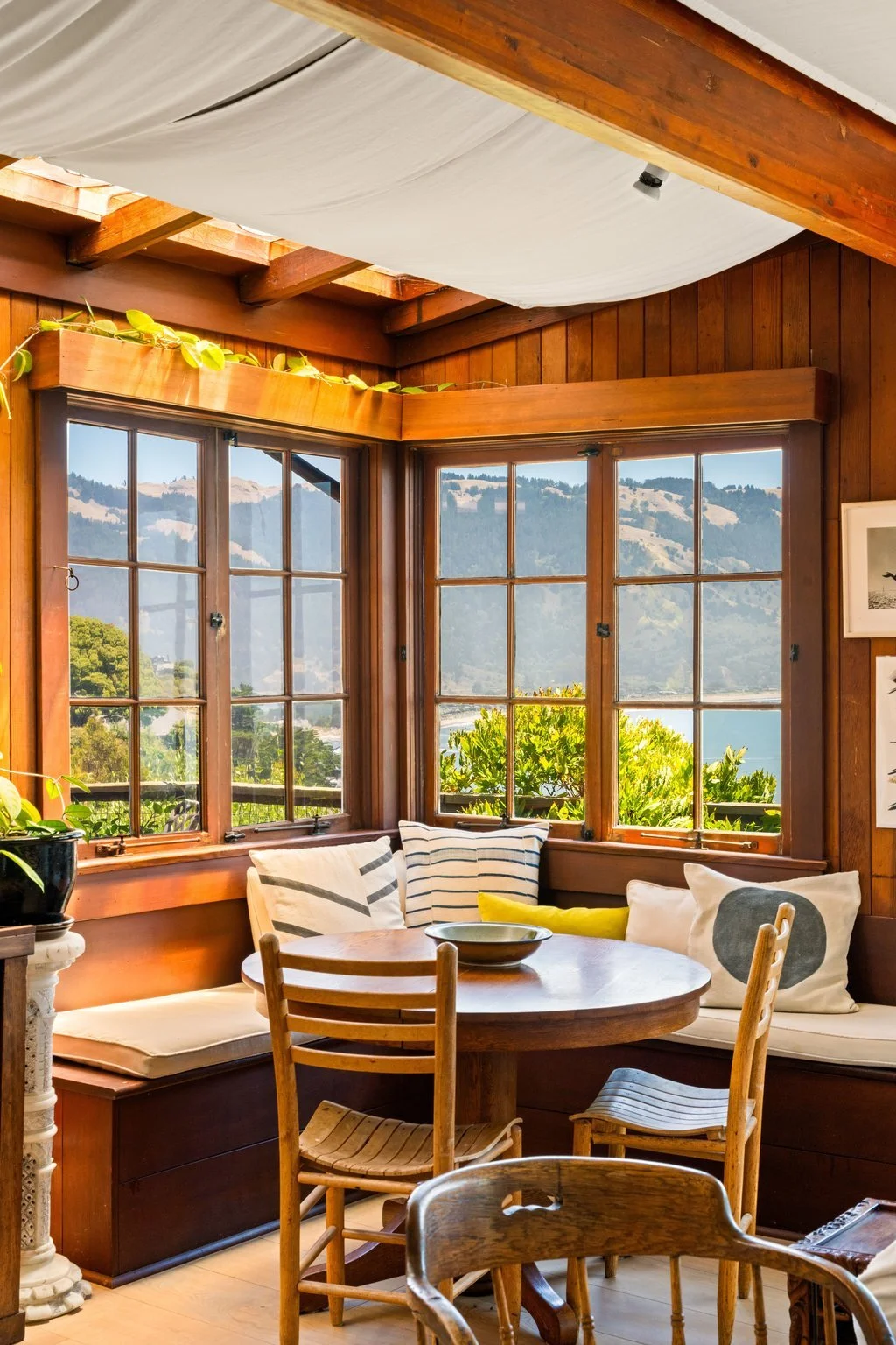 Cozy corner of a wooden room with large windows showing hills and water outside, a round wooden table with chairs, and cushions on a bench.