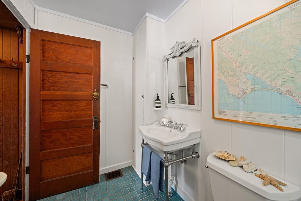 Small bathroom with white walls, a wooden door, a vintage white sink with a mirror above it, a toilet with seashell decorations, a framed map on the wall, and a small towel hanging on the sink.