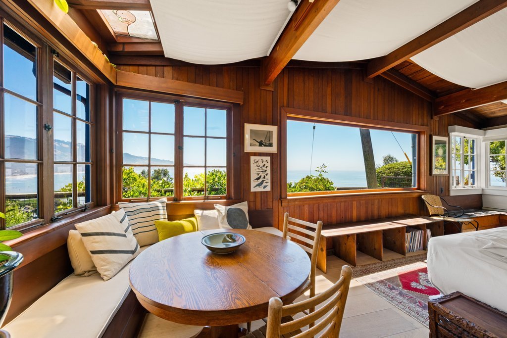 A cozy living space with large windows showing a scenic ocean view, wooden interior decor, a round wooden dining table with a bowl, a built-in bench with cushions, and a view of trees and water outside.