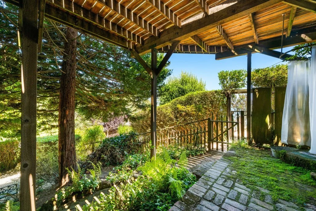 View of a backyard patio with a wooden roof, stone pathway, green plants, bushes, and trees, with a white curtain hanging on the right side and a wooden fence in the background.