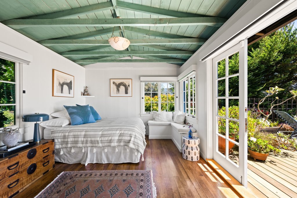 Bright bedroom with green painted vaulted ceiling, white walls, hardwood flooring, and a door leading to a sunlit outdoor deck with potted plants.