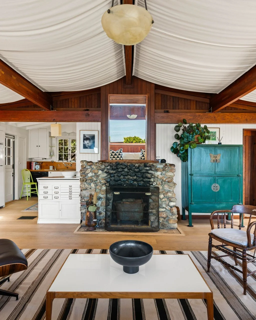 Interior of a living room with a stone fireplace, a white coffee table with a black bowl, a wooden armchair with a white cushion, and a turquoise cabinet, all under a white fabric ceiling with exposed wooden beams.