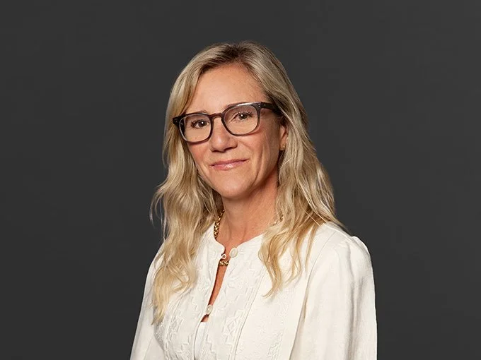 A woman with long blonde hair, wearing glasses and a white blouse, standing against a gray background.