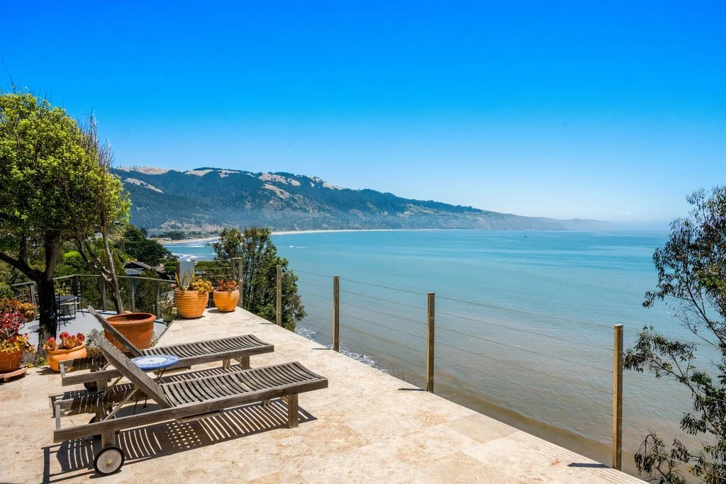 A coastal patio with lounge chairs, potted plants, and scenic ocean and mountain view under a clear blue sky.