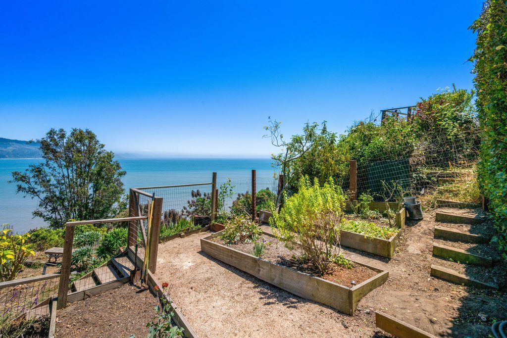 View of a garden with wooden raised beds, a gravel pathway, and a set of stairs leading up on the right, overlooking a body of water with trees and land in the background under a clear blue sky.