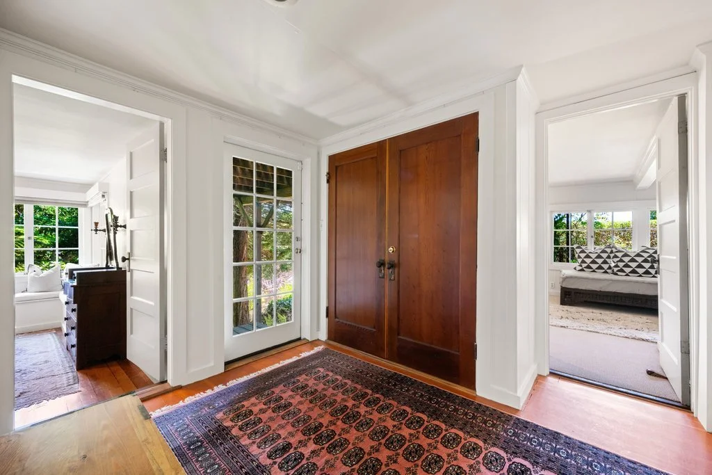 View of an entryway with a wooden cabinet, a glass door, and two open rooms on either side, featuring white walls, large windows, and furniture with decorative pillows.