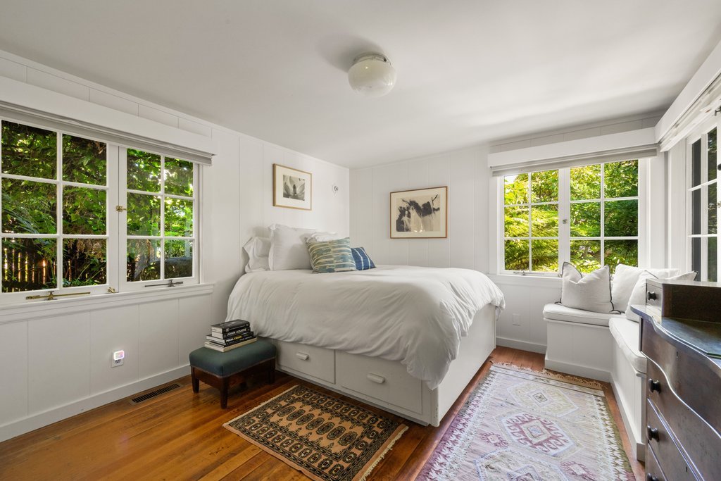 Bright bedroom with white walls, large windows showing greenery outside, a bed with white bedding, decorative pillows, a small upholstered stool with books, framed artwork, and a wooden floor with area rugs.