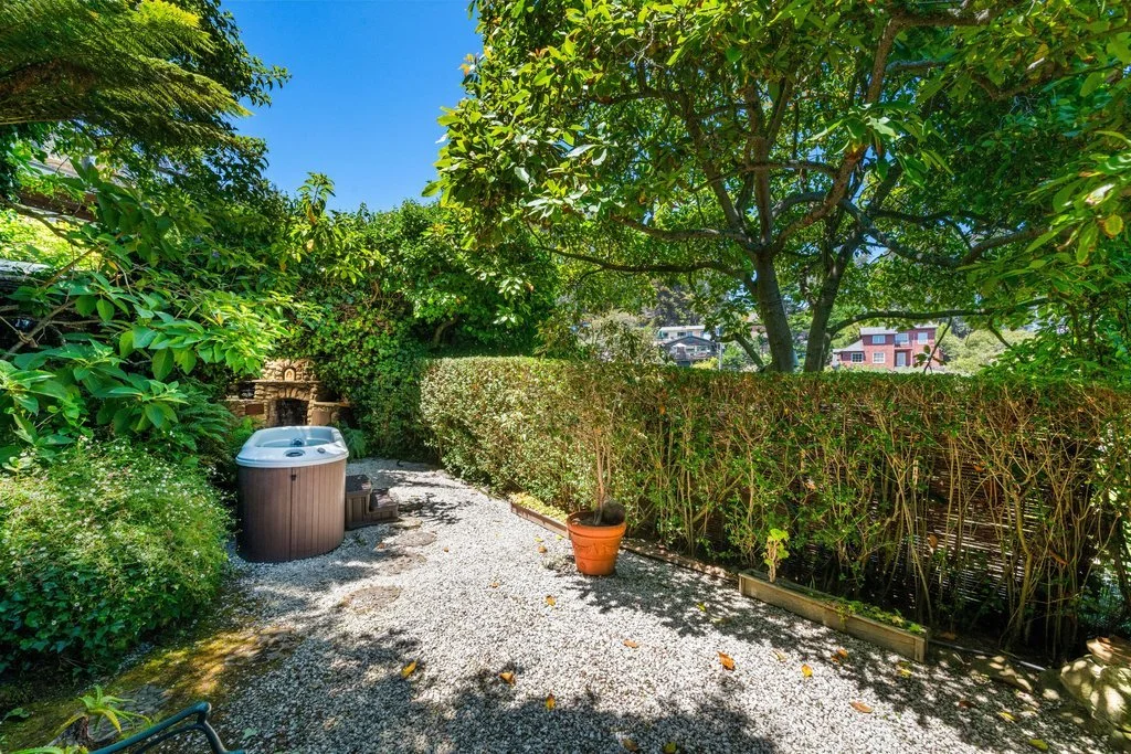 A backyard garden with green shrubs, a large tree, and a gravel pathway, featuring a hot tub, a potted plant, and a small outdoor fireplace.