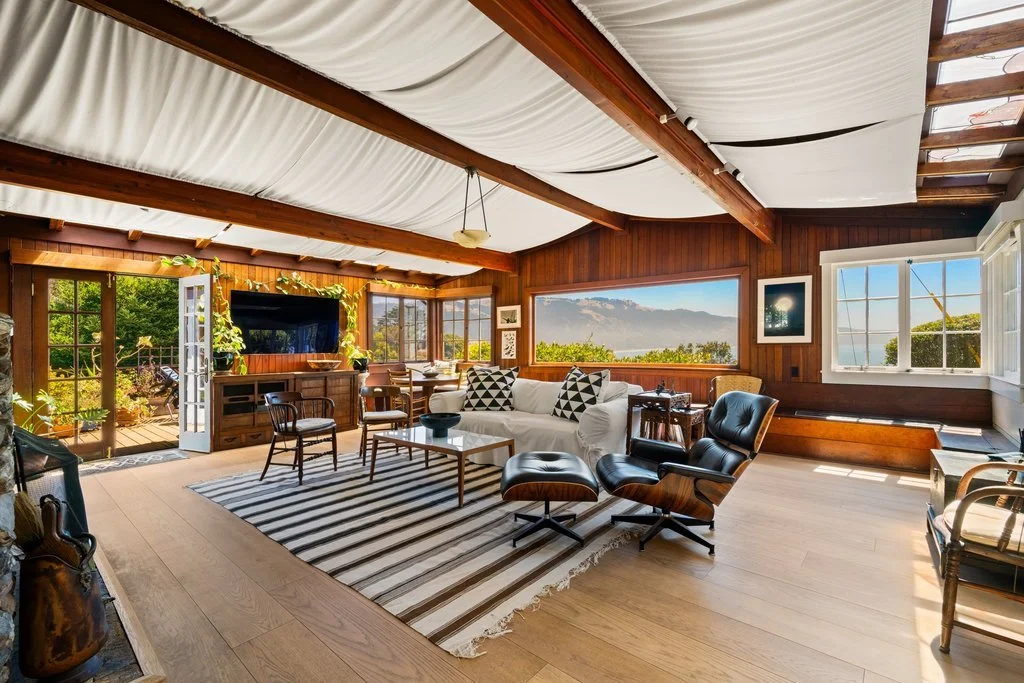 Sunlit living room with wooden paneling, large windows showing greenery, white sofa, black lounge chair with ottoman, striped rug, and a view of the sky and landscape.