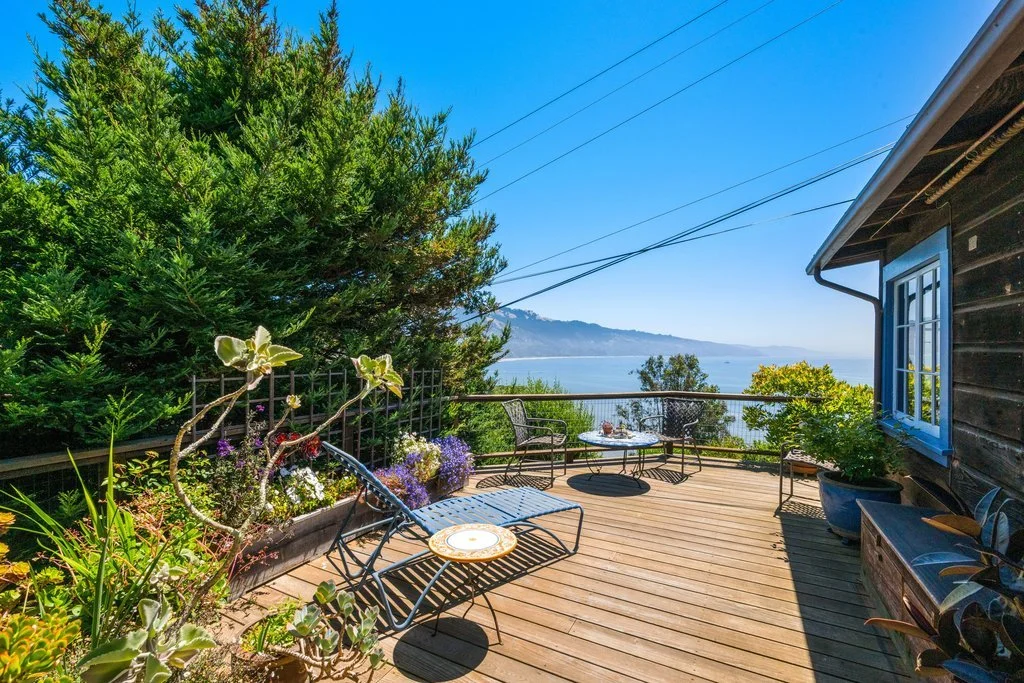 A wooden balcony with outdoor furniture overlooking the ocean and mountains, surrounded by potted plants and greenery.