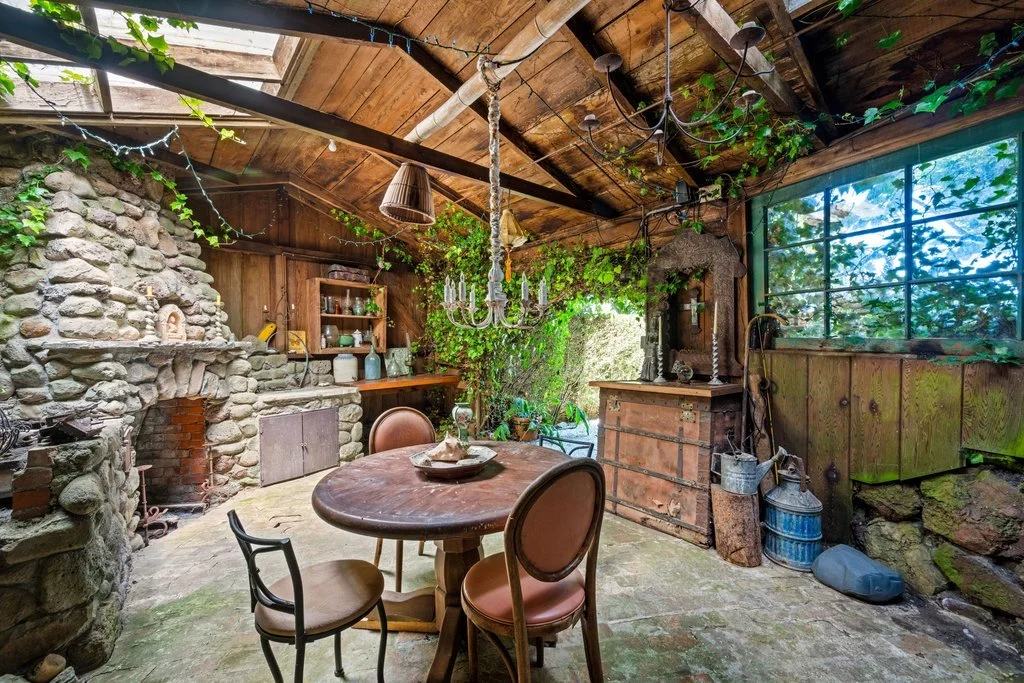 Rustic indoor space with a stone fireplace, wooden shelves with dishes, a wooden table with four chairs, greenery on the walls, and a large window with outside view.