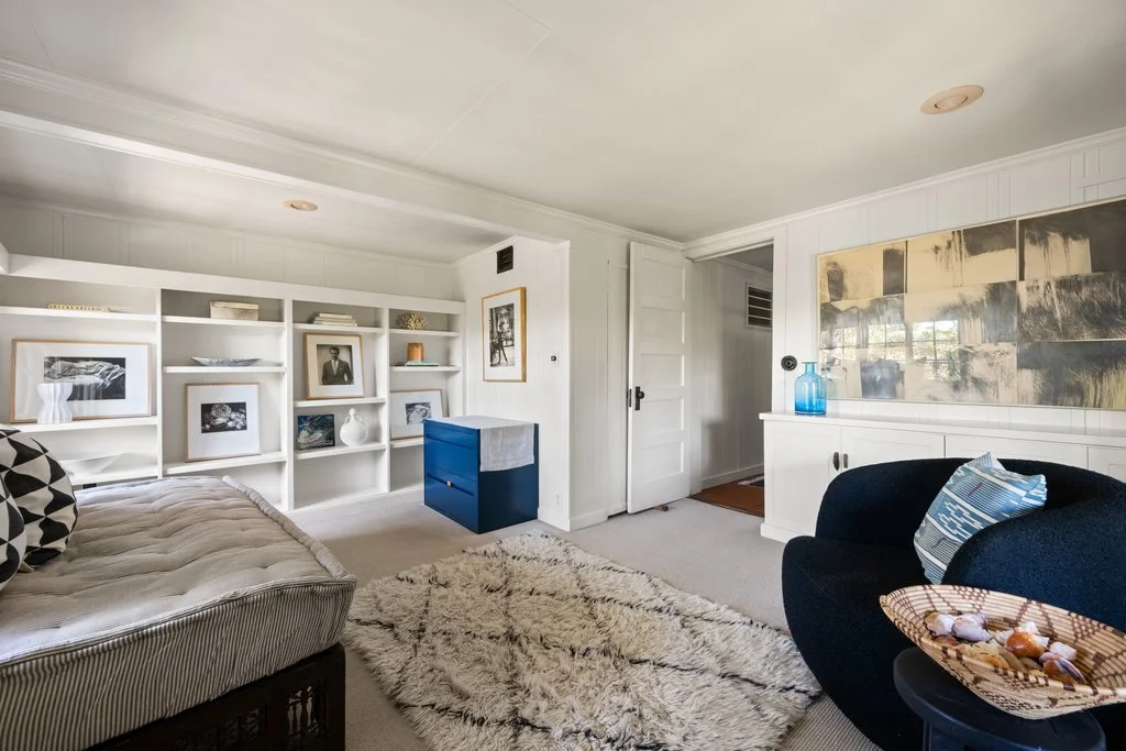 Living room with white built-in shelves, framed black-and-white artwork, a cream tufted sofa, a blue accent table, a shaggy beige rug, a navy armchair with a striped pillow, and a large abstract painting on the wall with decorative items including a 