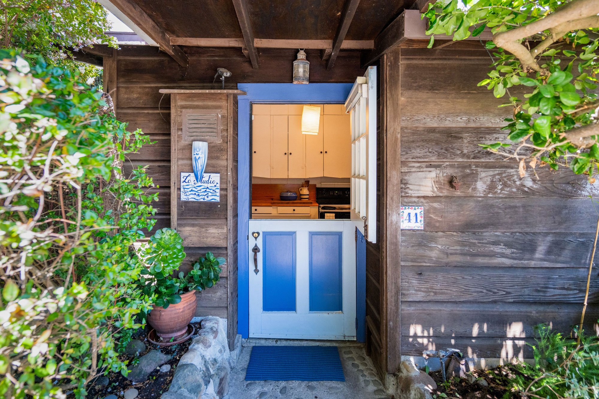 A wooden doorway with a blue door in a rustic wooden cabin. There are green plants and a potted plant on the left. The door is partially open, revealing a warmly lit interior kitchen with yellow cabinets and a paper lantern light. The cabin's wall is