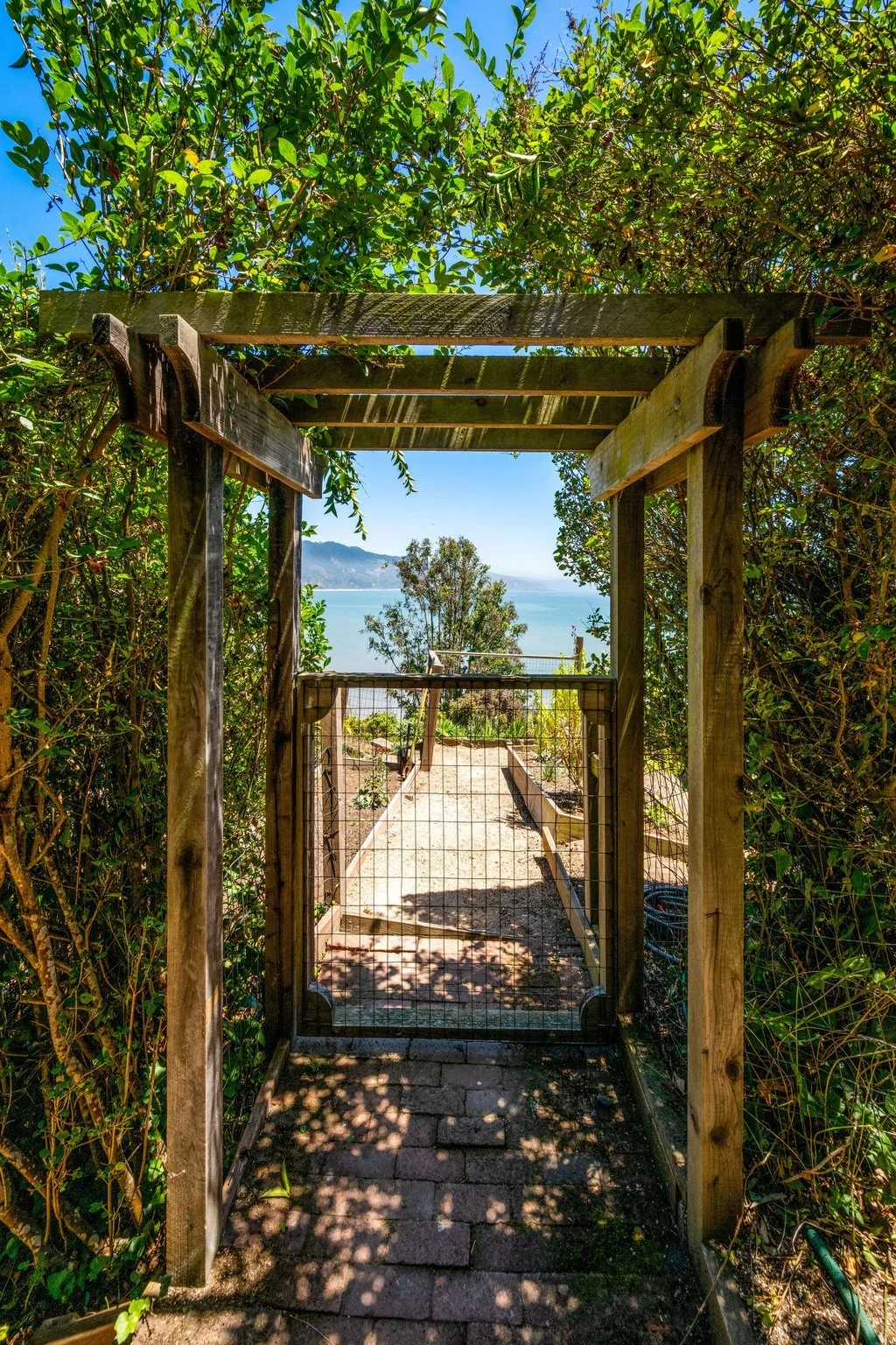 A wooden gate in a lush foliage entrance leads to a scenic pathway with a view of a body of water and mountains in the distance, under a clear blue sky.