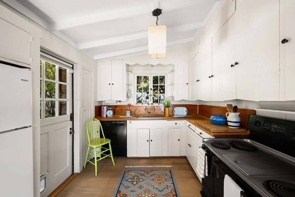 Kitchen with white cabinets, a wooden countertop, a black stove, white and blue dishes, a green chair, and a window with greenery outside.