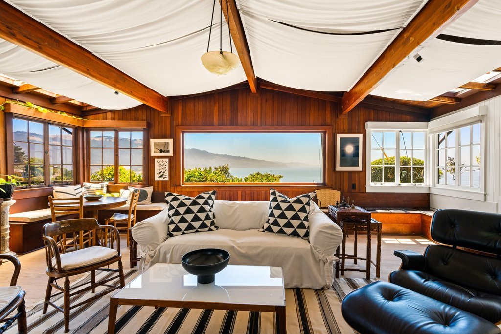 Living room with large windows, wooden ceiling beams, and ocean view in the background. Contains a white sofa with black and white geometric pillows, a black leather chair, a white coffee table, and a dining area with a round table and chairs.