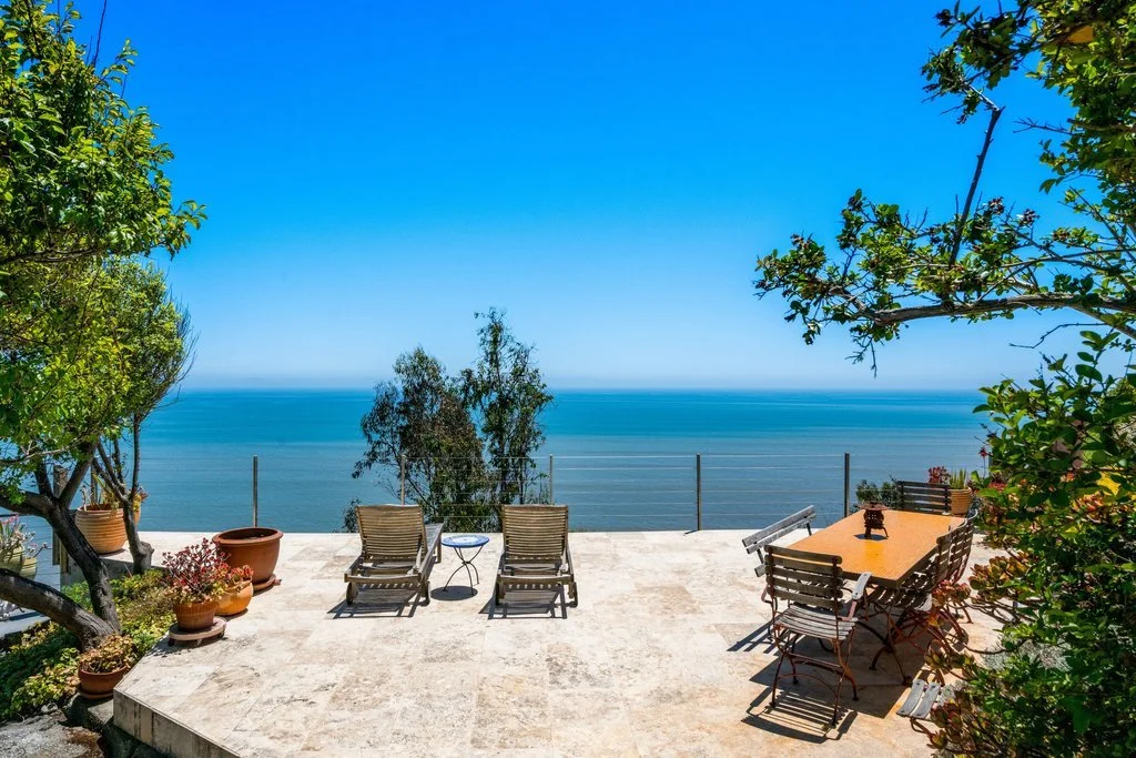 Patio with outdoor furniture overlooking the ocean, enclosed by a glass barrier, surrounded by green plants and trees.