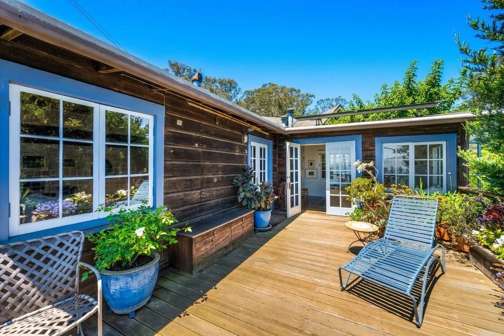 A wooden deck outdoor patio with outdoor furniture, potted plants, and a view of a house with large windows and a glass door, surrounded by greenery and trees under a bright blue sky.