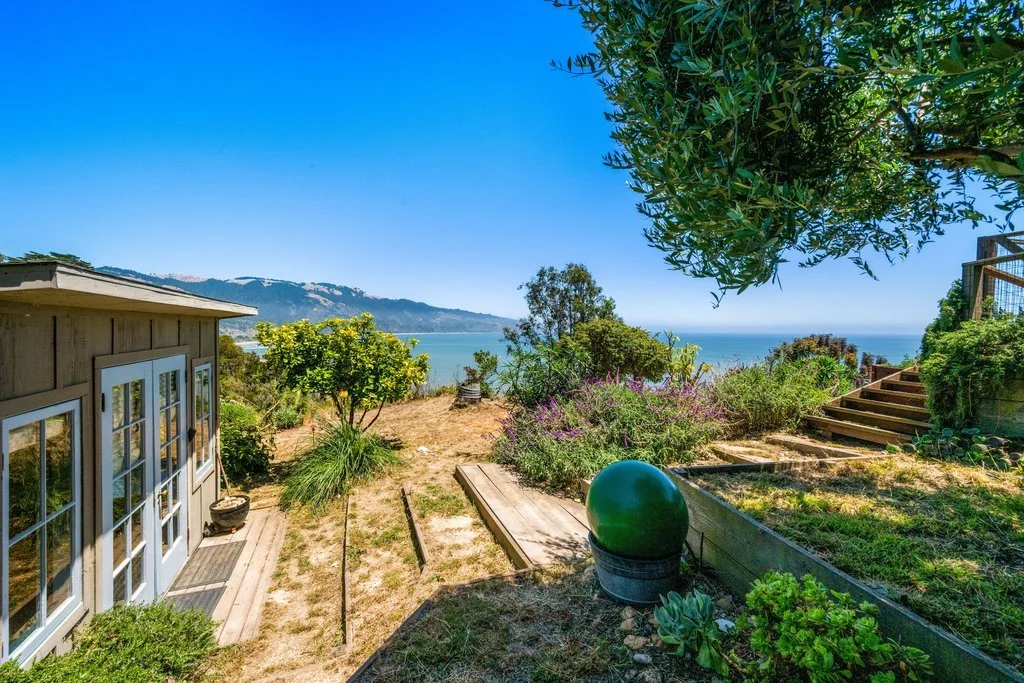 View of a coastal backyard with steps, plants, trees, and a view of the ocean and distant hills on a bright, sunny day.