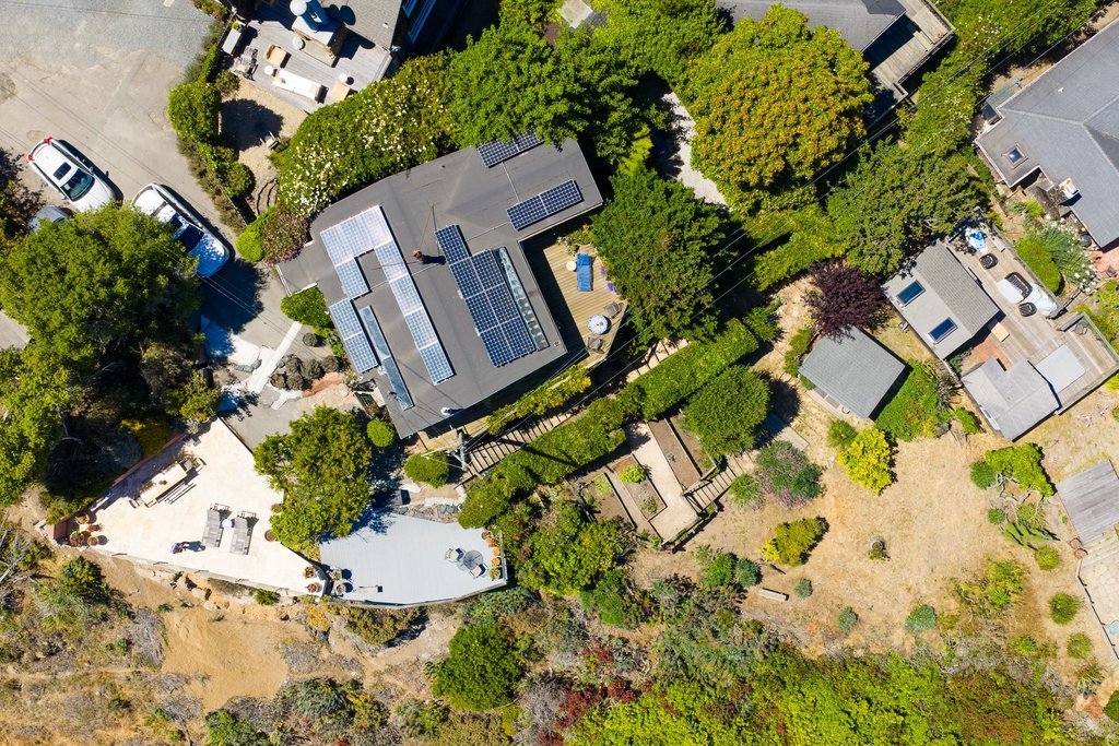 An aerial view of a residential backyard with trees, a house with solar panels on the roof, a deck, and a surrounding fence.
