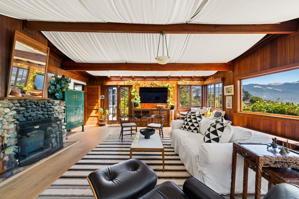 A cozy living room with wooden walls and large windows showing a mountain view. It has a white couch with black-and-white pillows, a striped rug, a black leather chair, and a stone fireplace.