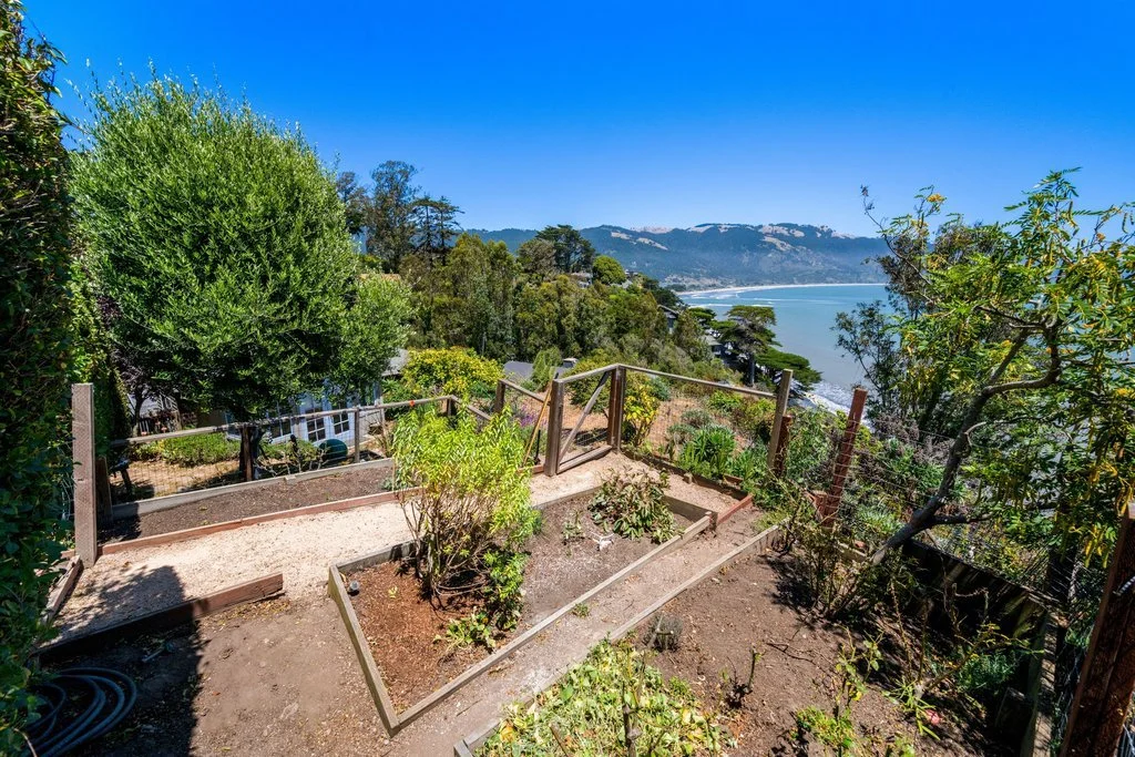 A backyard garden with raised planting beds, situated on a hillside overlooking the ocean and distant mountains under a clear blue sky.