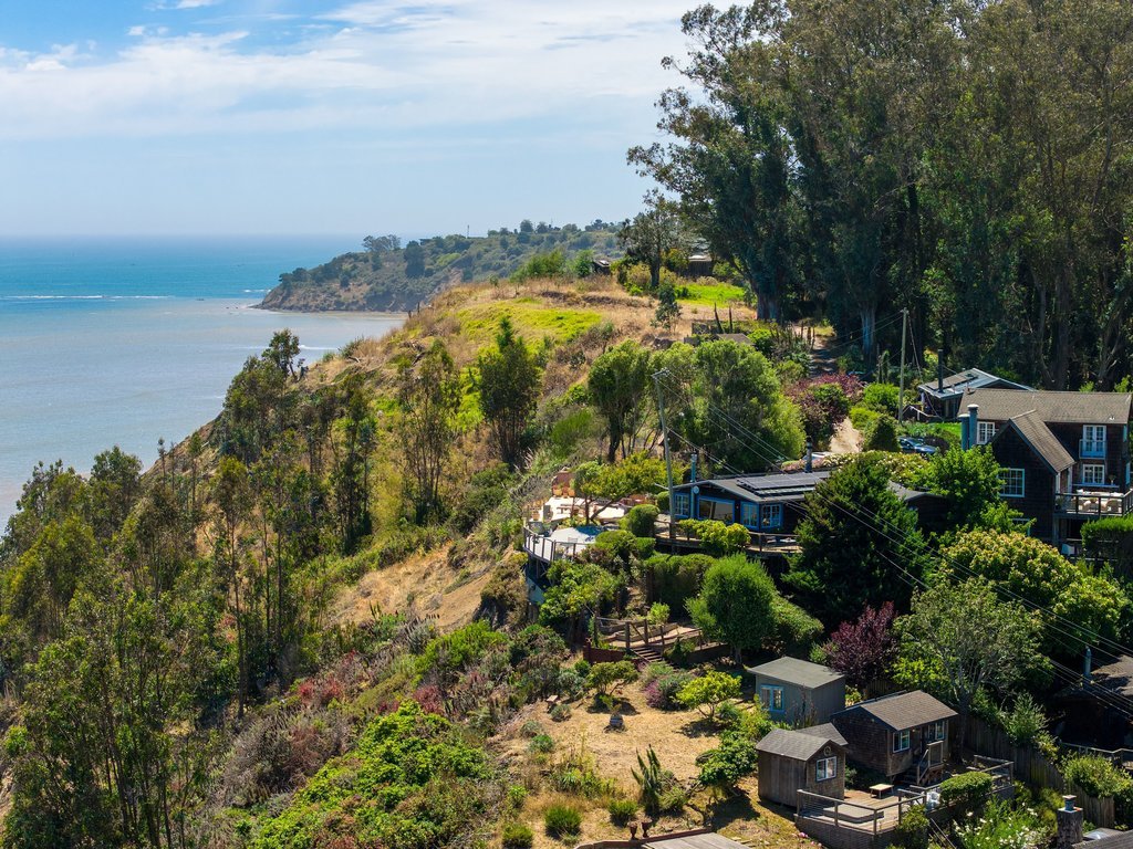 Houses and lush greenery on a hillside overlooking the ocean with a partly cloudy sky