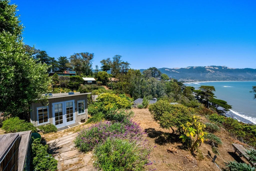 A coastal hillside garden with trees and shrubs overlooking the ocean and distant mountains on a sunny day.