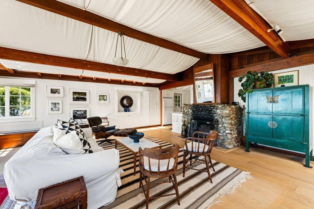Living room with white sofa, wooden chairs, stone fireplace, teal cabinet, framed artwork, and large windows with greenery outside.