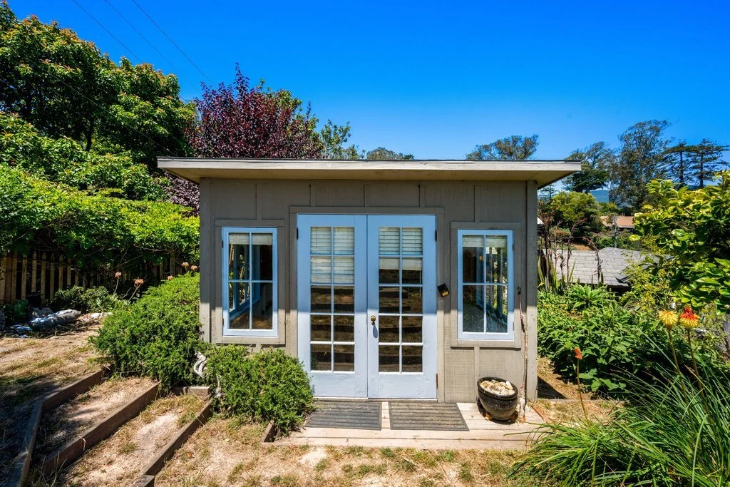Small garden shed with double glass doors and surrounding lush greenery under a clear blue sky.