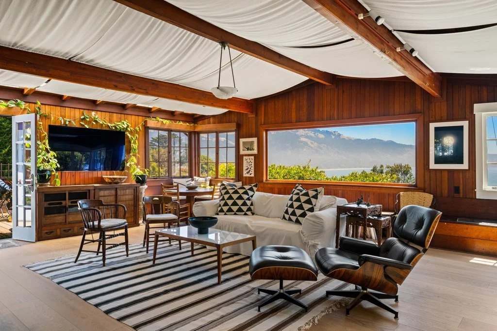 Living room with wooden walls and ceiling, featuring a large window and a glass door opening to a balcony, with a view of mountains and water. Contains a black leather chair and ottoman, white sofa with black and white pillows, striped rug, televisio