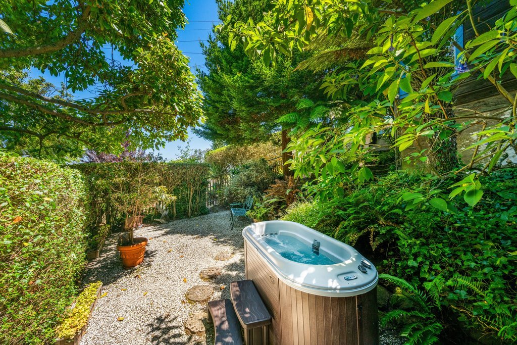A backyard with a hot tub, surrounded by lush green trees, bushes, and potted plants, with a gravel pathway and a garden bench in the background.