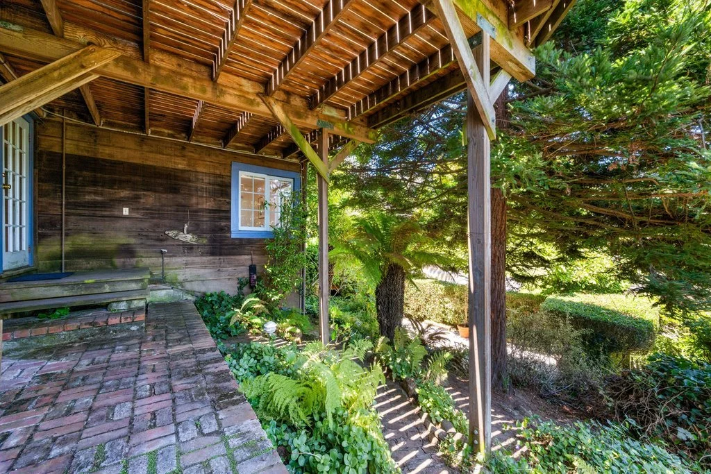 Back porch with brick flooring, wooden wall with a small window, and lush green trees and plants providing shade.