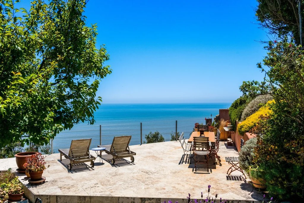 Outdoor terrace overlooking the ocean with lounge chairs and a dining area, surrounded by green plants and trees under a bright blue sky.
