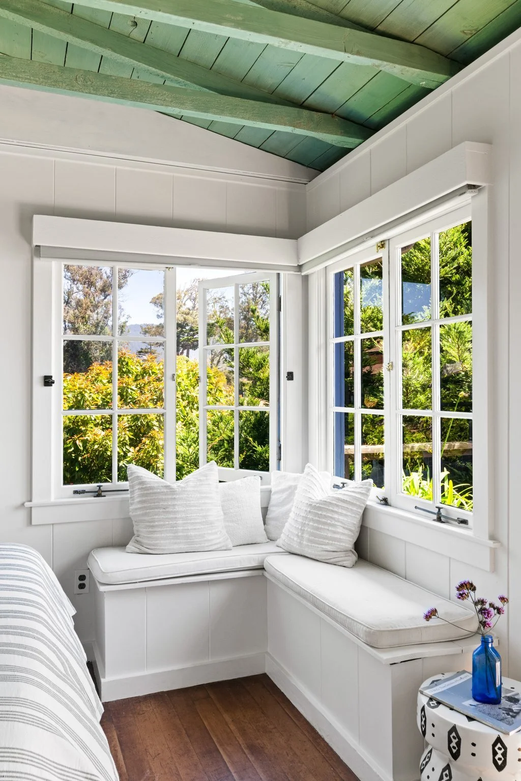 Cozy white sunroom with large windows showing lush greenery outside, white built-in window seat with white cushions, and a small side table with a blue vase containing purple flowers.