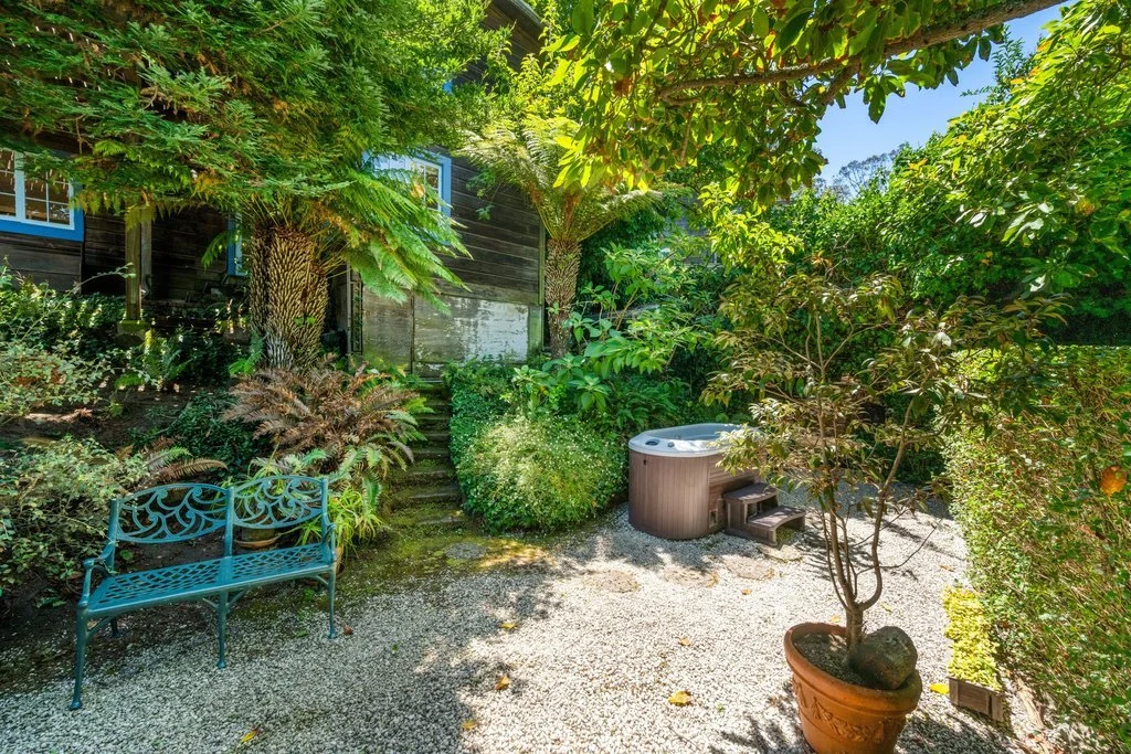 A lush backyard garden with various plants, a turquoise metal bench, a small hot tub, and potted trees under a canopy of leaves.