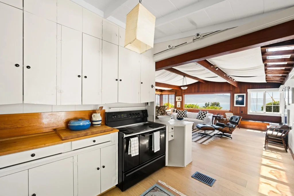 A cozy living room combined with a kitchen area, featuring white cabinetry, a black stove, and a view of the ocean through large windows, with wooden accents and a ceiling draped with white fabric.