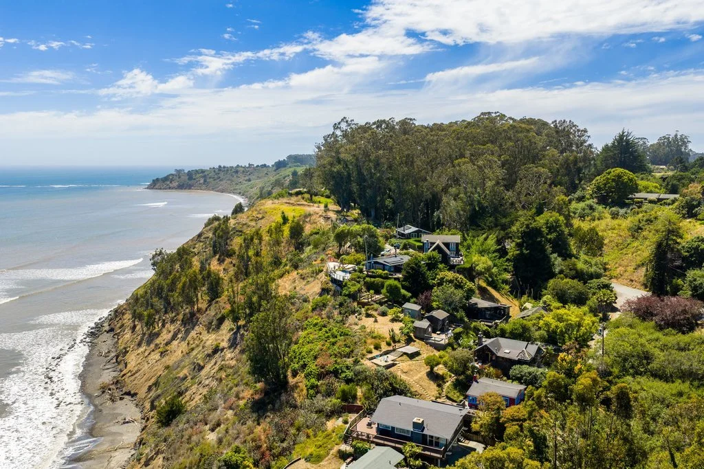 Cliffside homes overlooking the ocean with lush trees and sky in the background.