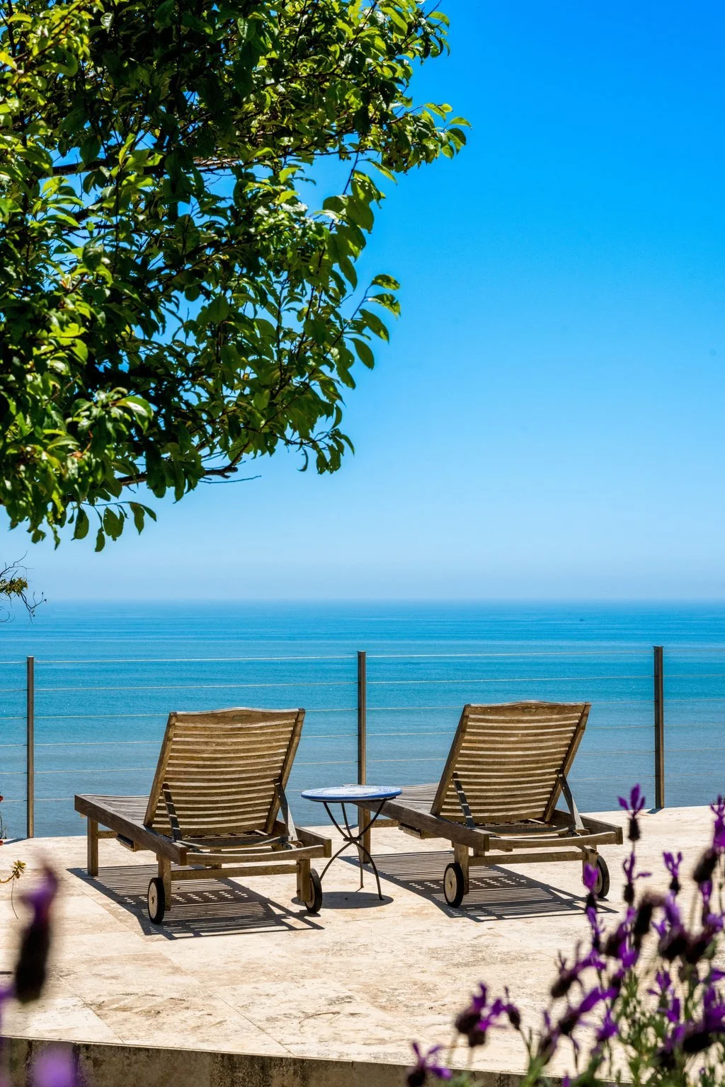 Two wooden lounge chairs on a terrace overlooking the ocean, with a small round table between them and purple flowers in the foreground. A large leafy tree is on the left, and the sky is clear and blue.