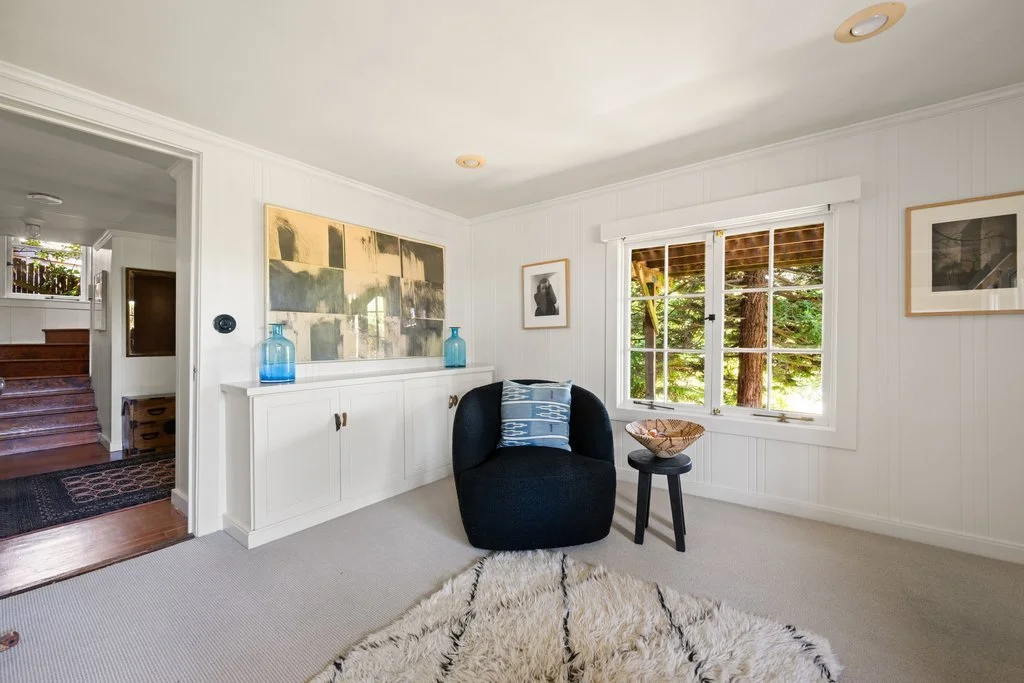 A cozy living room with white paneled walls, a black armchair with a blue pillow, a small black side table with a decorative bowl, a window with a view of greenery, and framed artwork on the walls. There's a white built-in cabinet with blue vases and