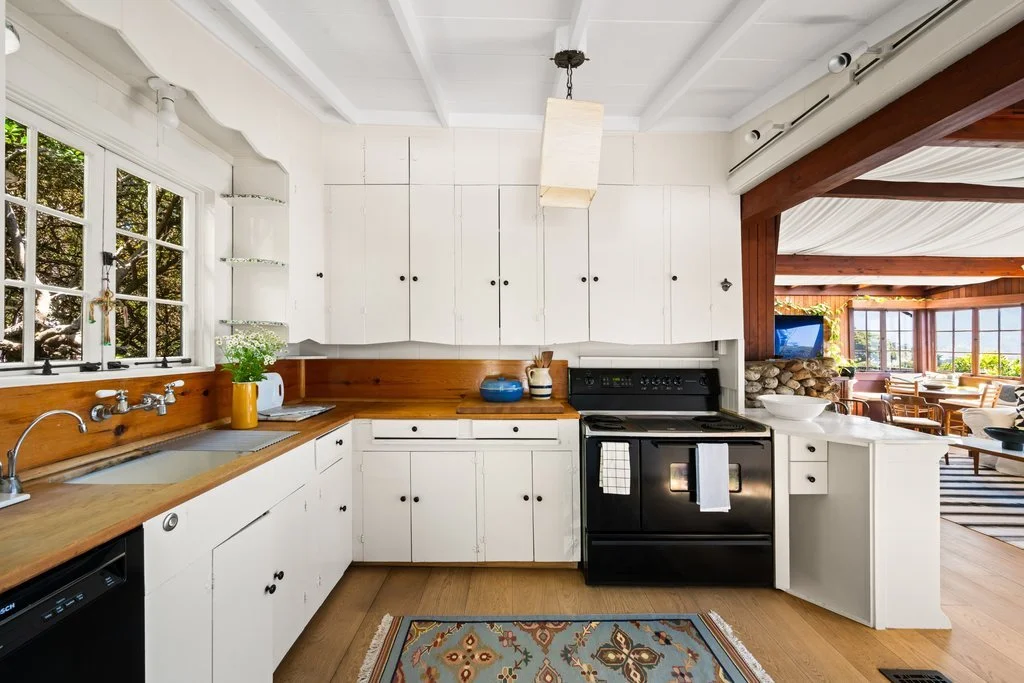 Kitchen with white cabinets, wooden countertops, black stove, and large window overlooking a garden.