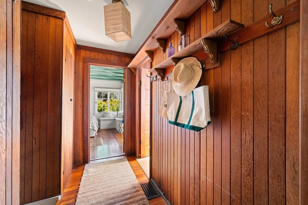 Wood-paneled hallway with hooks holding a straw hat and tote bag, leading to a bedroom with large window and white furniture.