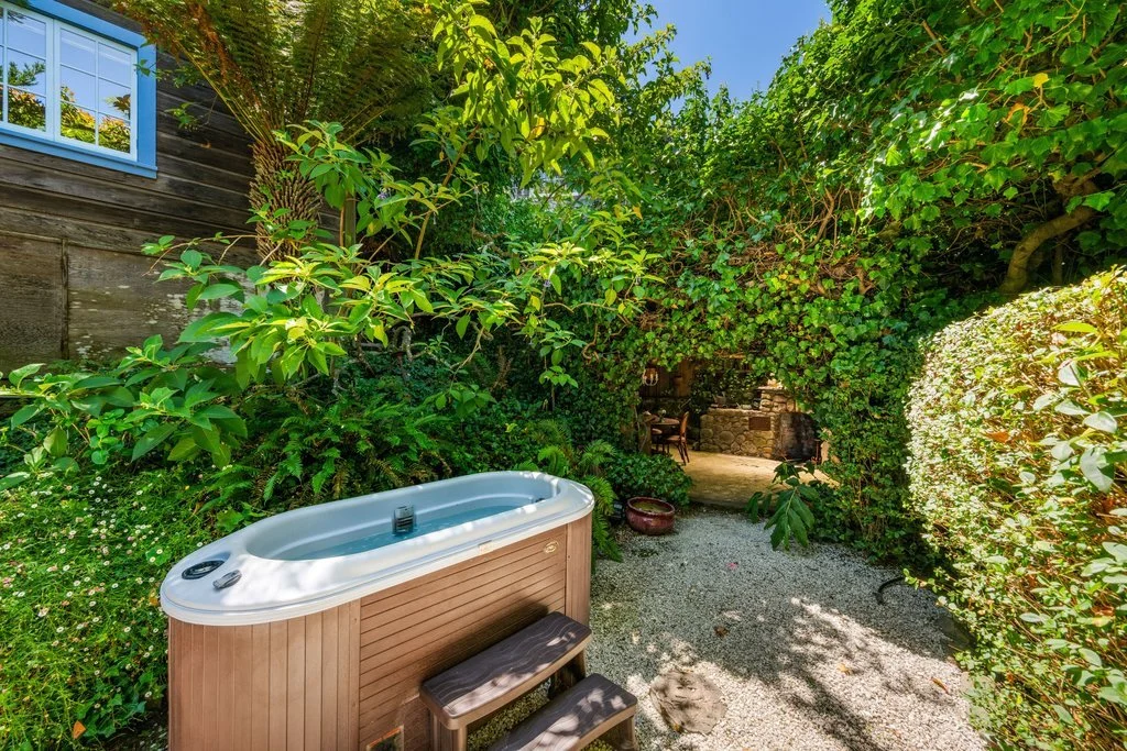 Outdoor backyard with hot tub, lush green trees and bushes, stone pathway, and a cozy seating area with a fireplace in the background.