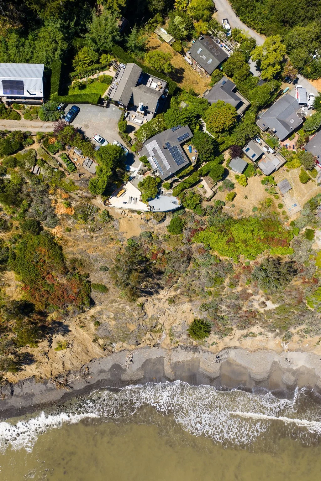 Aerial view of coastal residential area with houses, trees, and a beach with waves.