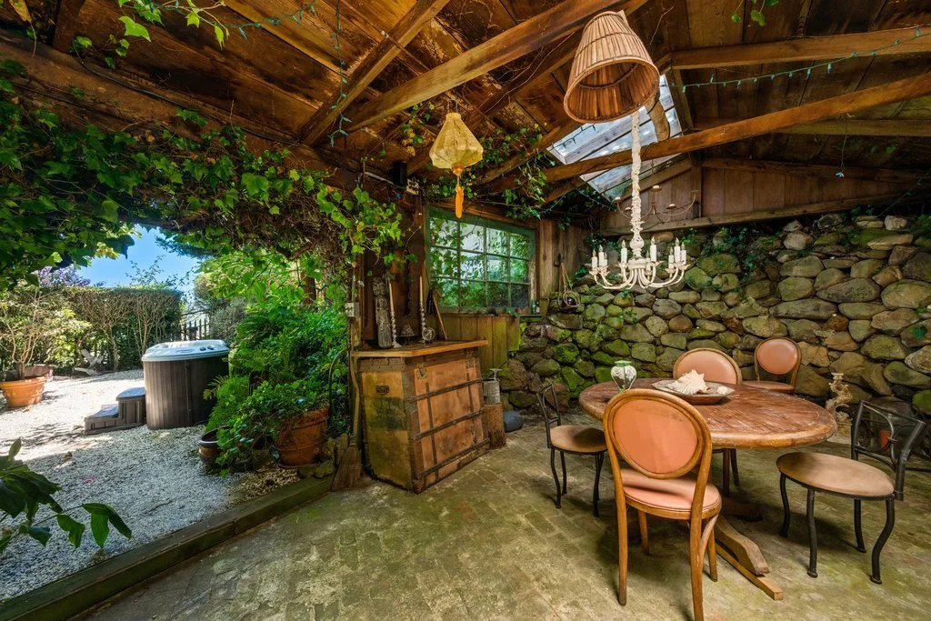 Indoor rustic dining area with a stone wall, round wooden table, pink upholstered chairs, hanging chandelier with a lampshade, and a large window leading to an outdoor garden with gravel ground, plants, and a hot tub.