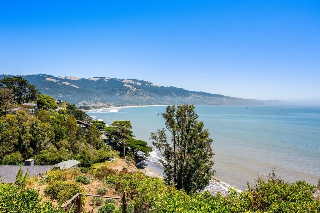 Coastal landscape with a clear blue sky, overlooking a beach, green trees, and houses on a hillside.