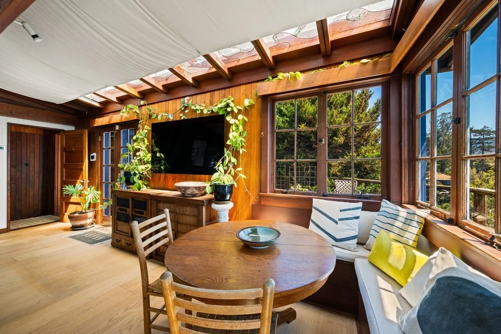 Cozy sunroom with large windows, wooden walls, a round dining table, and a built-in bench with cushions. A TV is mounted on the wooden wall, surrounded by plants, and there is a potted plant near the door.