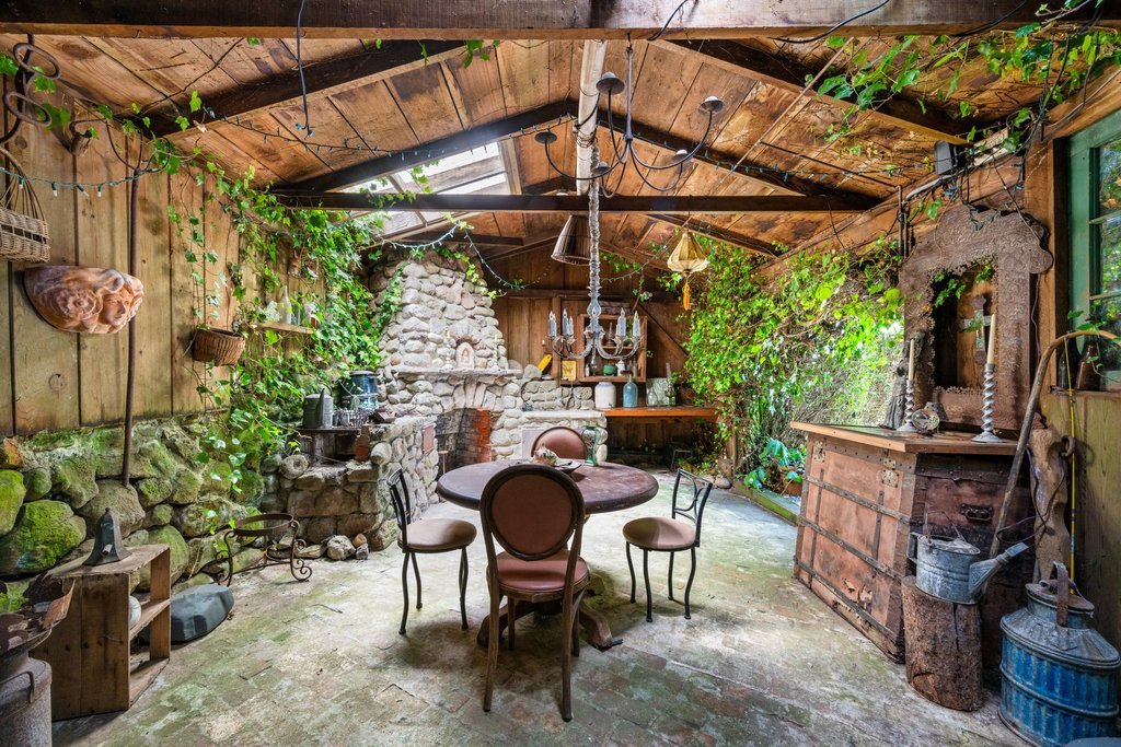 Rustic indoor space with wooden walls and ceiling, green plants, a stone fireplace, round dining table with four chairs, and various decorations and shelves.