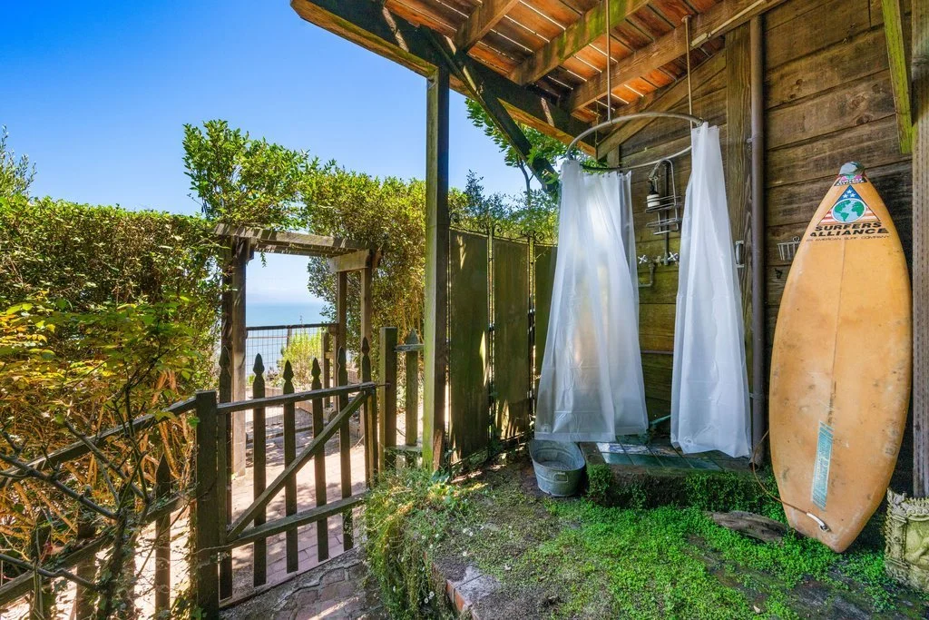 Outdoor shower with white curtains next to a surfboard, overlooking the ocean through a wooden gate and lush greenery.